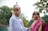 Two children are dressed in traditional attire, the boy in a white kurta with a headband and red markings on his forehead, while the girl is wearing a pink and gold sari, accessorized with jewelry including a forehead ornament. They are outdoors with trees in the background.