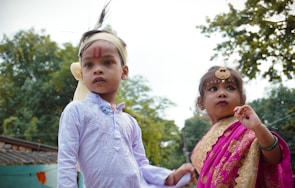 Two children are dressed in traditional attire, the boy in a white kurta with a headband and red markings on his forehead, while the girl is wearing a pink and gold sari, accessorized with jewelry including a forehead ornament. They are outdoors with trees in the background.