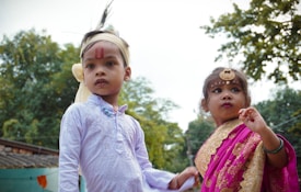 Two children are dressed in traditional attire, the boy in a white kurta with a headband and red markings on his forehead, while the girl is wearing a pink and gold sari, accessorized with jewelry including a forehead ornament. They are outdoors with trees in the background.