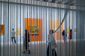A modern art gallery with people walking and observing paintings on the walls, viewed through vertical metal or string elements. The space is well-lit with large, colorful canvases mounted against white walls.
