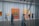 A modern art gallery with people walking and observing paintings on the walls, viewed through vertical metal or string elements. The space is well-lit with large, colorful canvases mounted against white walls.