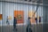 A modern art gallery with people walking and observing paintings on the walls, viewed through vertical metal or string elements. The space is well-lit with large, colorful canvases mounted against white walls.