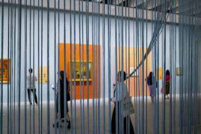 A modern art gallery with people walking and observing paintings on the walls, viewed through vertical metal or string elements. The space is well-lit with large, colorful canvases mounted against white walls.