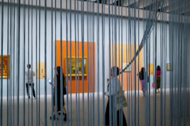 A modern art gallery with people walking and observing paintings on the walls, viewed through vertical metal or string elements. The space is well-lit with large, colorful canvases mounted against white walls.