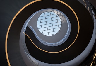 A spiraling architectural structure reminiscent of a helical staircase viewed from below, showcasing a grid-patterned skylight at the top. Soft ambient lighting adds a modern and futuristic element.