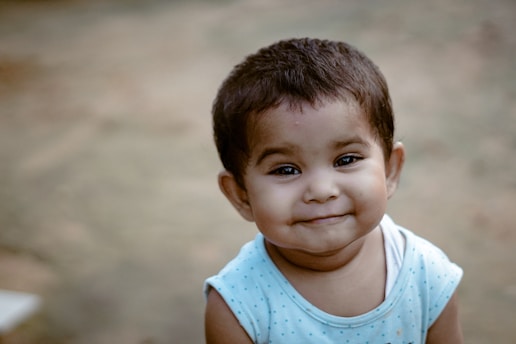 A young child with short hair smiles warmly, wearing a light blue sleeveless shirt with small polka dots. The background is a blurred earthy tone, giving the image a soft focus on the child.