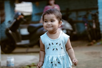 A young child with short hair is wearing a light blue dress with floral patterns. The child is standing outdoors on a slightly blurred background that includes a scooter, some bicycles, and a person in casual clothing. The child has a cheerful expression and is positioned slightly off-center.