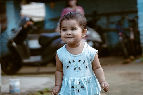 A young child with short hair is wearing a light blue dress with floral patterns. The child is standing outdoors on a slightly blurred background that includes a scooter, some bicycles, and a person in casual clothing. The child has a cheerful expression and is positioned slightly off-center.
