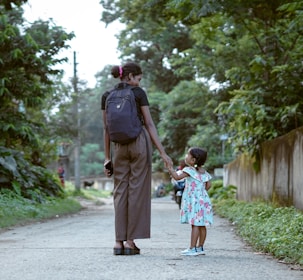A woman and a young girl walk hand in hand down a tree-lined path, surrounded by lush greenery. The woman carries a backpack and holds a phone, while the girl wears a floral dress and sneakers, gazing upwards.