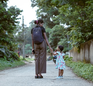 A woman and a young girl walk hand in hand down a tree-lined path, surrounded by lush greenery. The woman carries a backpack and holds a phone, while the girl wears a floral dress and sneakers, gazing upwards.