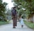 A woman and a young girl walk hand in hand down a tree-lined path, surrounded by lush greenery. The woman carries a backpack and holds a phone, while the girl wears a floral dress and sneakers, gazing upwards.