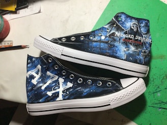 A pair of custom-painted high-top sneakers featuring artwork themed around a music band. One shoe displays a grim reaper figure and the band's name with the word 'Nightmare'. The other shoe showcases the letters 'A7X' in a bold style. The background combines dark and mystical elements with shades of blue and white to create a celestial effect.
