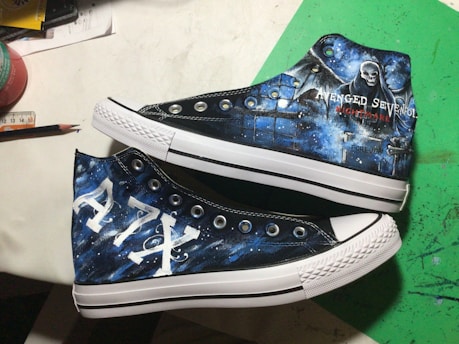 A pair of custom-painted high-top sneakers featuring artwork themed around a music band. One shoe displays a grim reaper figure and the band's name with the word 'Nightmare'. The other shoe showcases the letters 'A7X' in a bold style. The background combines dark and mystical elements with shades of blue and white to create a celestial effect.