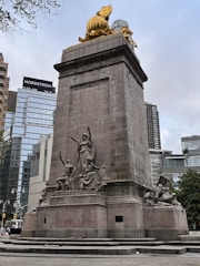 A large stone monument adorned with several detailed sculptures, including seated and standing figures. Atop the monument is a shiny, golden statue, set against a backdrop of modern skyscrapers with glass facades. Trees and urban elements are visible in the surroundings.