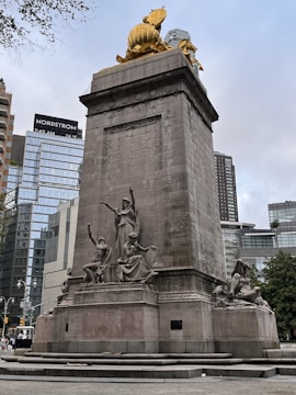 A large stone monument adorned with several detailed sculptures, including seated and standing figures. Atop the monument is a shiny, golden statue, set against a backdrop of modern skyscrapers with glass facades. Trees and urban elements are visible in the surroundings.