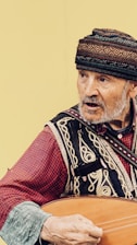An elderly man is wearing traditional attire with intricate patterns and a colorful hat. He appears to be playing a musical instrument, possibly a stringed one, with his right hand. The background is a plain, light color.