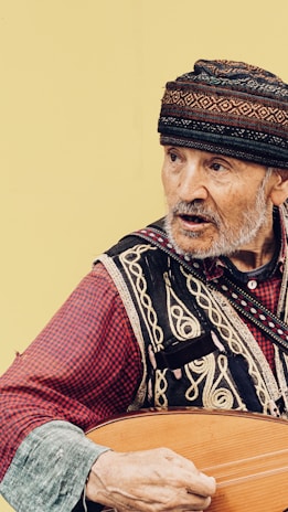 An elderly man is wearing traditional attire with intricate patterns and a colorful hat. He appears to be playing a musical instrument, possibly a stringed one, with his right hand. The background is a plain, light color.