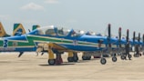 A row of blue and yellow propeller aircraft parked on a tarmac with visible markings and insignias. The planes are arranged neatly in a line, with military personnel standing beside them. The sky is clear with a few clouds visible.