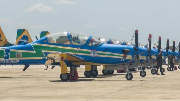 A row of blue and yellow propeller aircraft parked on a tarmac with visible markings and insignias. The planes are arranged neatly in a line, with military personnel standing beside them. The sky is clear with a few clouds visible.