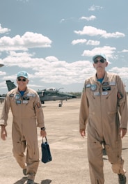 Two individuals dressed in pilot uniforms walk on an airfield with a military aircraft in the background. The sky is bright with scattered clouds, creating a serene atmosphere.