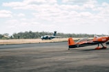 Two airplanes are situated on an airfield. In the foreground, a small orange and black aerobatic aircraft is stationary on the tarmac. In the background, a large green military cargo plane is taxiing on the runway. The airfield is bordered by a line of trees, and the sky is partly cloudy.