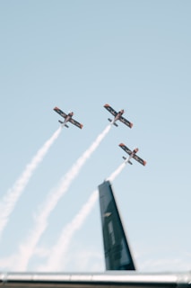 Three aerobatic airplanes with trailing smoke are seen flying in a formation against a clear blue sky. The focus is on the planes, and there is a faint outline of a large black structure at the bottom of the image, likely part of an aircraft.