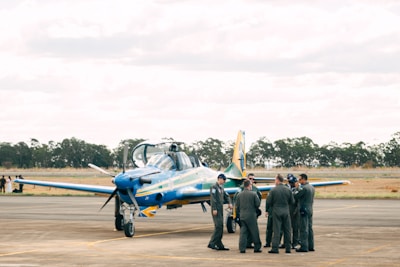 A group of people in military or aviation uniforms stand together on an airfield near a blue and yellow aircraft. The setting appears to be outdoors with a backdrop of trees and an overcast sky.