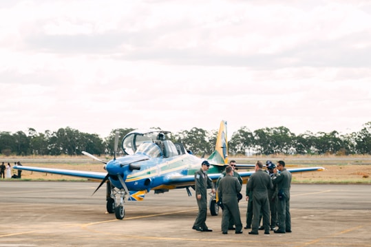 A group of people in military or aviation uniforms stand together on an airfield near a blue and yellow aircraft. The setting appears to be outdoors with a backdrop of trees and an overcast sky.