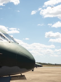 The image depicts a close-up view of a military aircraft on an airfield, with a clear blue sky and scattered white clouds in the background. Another aircraft is visible in the distance, and there seems to be a lone parachutist descending from above.