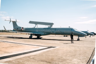 A military aircraft on a tarmac with a person standing in front of it. The aircraft has 'Força Aérea Brasileira' written on its side, indicating it belongs to the Brazilian Air Force. The sky is clear with a few clouds, and the environment is open and expansive.