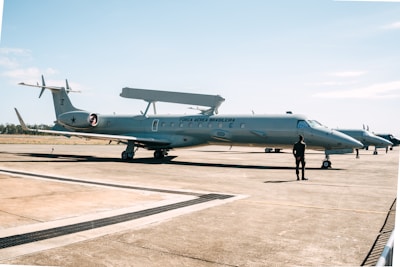 A military aircraft on a tarmac with a person standing in front of it. The aircraft has 'Força Aérea Brasileira' written on its side, indicating it belongs to the Brazilian Air Force. The sky is clear with a few clouds, and the environment is open and expansive.