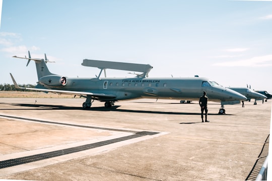 A military aircraft on a tarmac with a person standing in front of it. The aircraft has 'Força Aérea Brasileira' written on its side, indicating it belongs to the Brazilian Air Force. The sky is clear with a few clouds, and the environment is open and expansive.