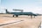 A military aircraft on a tarmac with a person standing in front of it. The aircraft has 'Força Aérea Brasileira' written on its side, indicating it belongs to the Brazilian Air Force. The sky is clear with a few clouds, and the environment is open and expansive.