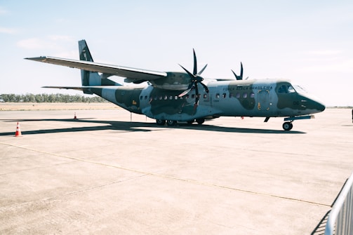 A military aircraft is parked on a concrete runway, with the words 'Força Aérea Brasileira' visible on its side. The plane has two propellers on each wing and is painted in a camouflage pattern. The sky is clear and the surrounding area appears empty, with only a few traffic cones visible on the ground.