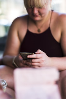 A person is sitting and holding a smartphone, with their eyes focused on the screen. The background is blurred, emphasizing the individual and the device in their hands. The person is wearing a dark tank top and has light blonde hair.