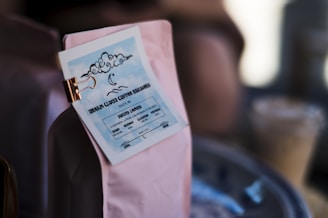 A close-up of a coffee bag with a label attached by a clip. The label features a cloud illustration with a crescent moon and wavy lines, reading 'Urban Cloud Coffee Roasters' and location details in stylized text. The image is softly lit, with some blurred objects in the background.