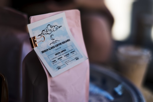 A close-up of a coffee bag with a label attached by a clip. The label features a cloud illustration with a crescent moon and wavy lines, reading 'Urban Cloud Coffee Roasters' and location details in stylized text. The image is softly lit, with some blurred objects in the background.