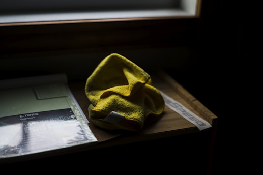 A yellow microfiber cloth rests on a wooden surface beside a pad of engineer's calculation paper. The scene is dimly lit by natural light coming from a nearby window, casting soft shadows. The wooden texture is visible, and there is a label reading 'speed' partially obscured by the cloth.