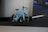 A small model of a bicycle with a blue frame and black accents is placed on a dark shelf. The bicycle features a minimalist design with two large, metallic, circular wheels. Adjacent to it is a poster or picture partially visible, showing part of a blue car with a rear view and road markings.