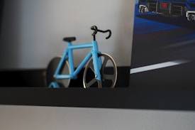 A small model of a bicycle with a blue frame and black accents is placed on a dark shelf. The bicycle features a minimalist design with two large, metallic, circular wheels. Adjacent to it is a poster or picture partially visible, showing part of a blue car with a rear view and road markings.