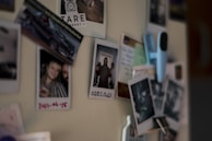 A collection of photographs and notes are attached to a beige surface, possibly a refrigerator. Some images show people smiling, and there are various items like a blue clip and cards in the vicinity.
