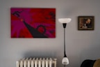 A modern, minimalist interior with a striking abstract painting in red and pink hues on the wall. The painting features geometric shapes and bold lines. To the right, a classic framed portrait depicts a contemplative figure. A sleek, black and white floor lamp stands between the paintings, and a white radiator is visible below.