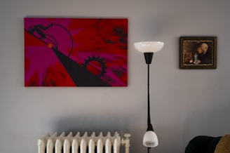 A modern, minimalist interior with a striking abstract painting in red and pink hues on the wall. The painting features geometric shapes and bold lines. To the right, a classic framed portrait depicts a contemplative figure. A sleek, black and white floor lamp stands between the paintings, and a white radiator is visible below.
