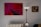 A modern, minimalist interior with a striking abstract painting in red and pink hues on the wall. The painting features geometric shapes and bold lines. To the right, a classic framed portrait depicts a contemplative figure. A sleek, black and white floor lamp stands between the paintings, and a white radiator is visible below.