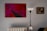 A modern, minimalist interior with a striking abstract painting in red and pink hues on the wall. The painting features geometric shapes and bold lines. To the right, a classic framed portrait depicts a contemplative figure. A sleek, black and white floor lamp stands between the paintings, and a white radiator is visible below.