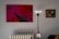 A modern, minimalist interior with a striking abstract painting in red and pink hues on the wall. The painting features geometric shapes and bold lines. To the right, a classic framed portrait depicts a contemplative figure. A sleek, black and white floor lamp stands between the paintings, and a white radiator is visible below.