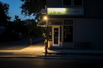 A dimly-lit street corner at dusk or early evening with a small commercial establishment, possibly a cafe or store, illuminated by a street light. The building features a sign with forest scenery and the name 'Canieen.' A construction cone and some other objects are visible on the sidewalk along with illuminated windows and door signs.
