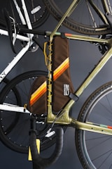 A close-up of a bicycle with a brown frame bag featuring orange and yellow stripes. The bag is attached to the main triangle of the bike's frame. The bike's handlebars, wheels, and other sections are visible, with another bike in the background. The image is set against a dark backdrop.
