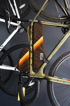 A close-up of a bicycle with a brown frame bag featuring orange and yellow stripes. The bag is attached to the main triangle of the bike's frame. The bike's handlebars, wheels, and other sections are visible, with another bike in the background. The image is set against a dark backdrop.
