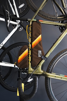 A close-up of a bicycle with a brown frame bag featuring orange and yellow stripes. The bag is attached to the main triangle of the bike's frame. The bike's handlebars, wheels, and other sections are visible, with another bike in the background. The image is set against a dark backdrop.